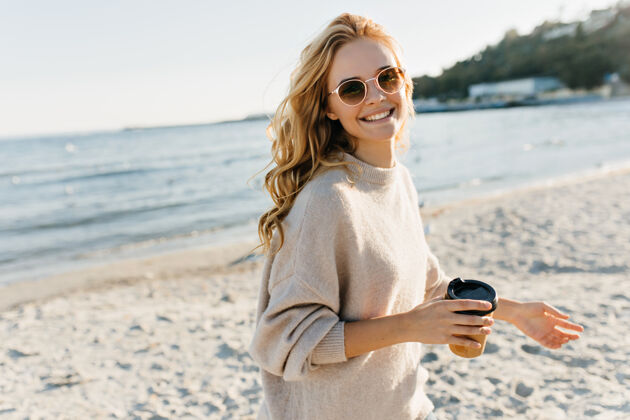 活动在沙滩上 一位迷人的盲人妇女手拿咖啡在寒冷的日子里 热情的戴着太阳镜的女模特在湖边摆姿势休闲茶自然