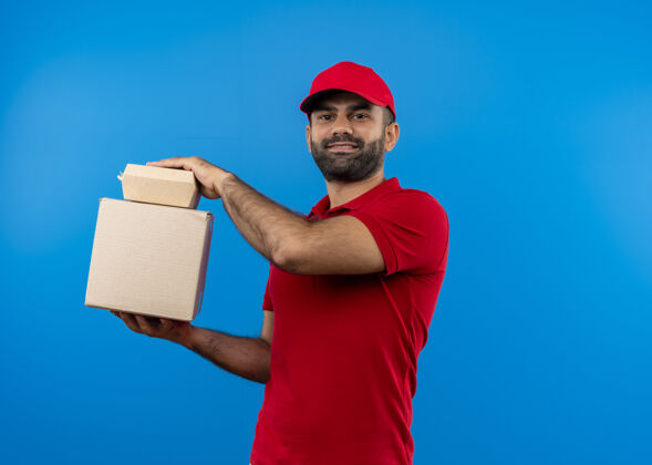 胡子满脸胡须 身穿红色制服 戴帽子的送货员站在蓝色的墙上 手里拿着盒子里的包裹 带着自信的微笑制服自信包装