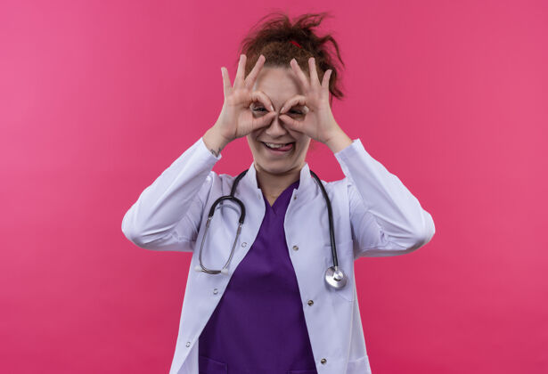 医生年轻的女医生 穿着白大褂 带听诊器 手指像望远镜一样 站在粉红色的墙上 伸出舌头 透过手指看 做着正常的手势女人穿过望远镜