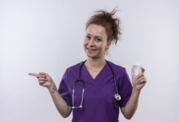 测试年轻的女医生穿着医疗制服 手持听诊器 手指指向一边 微笑着站在白色的墙上穿听诊器手指