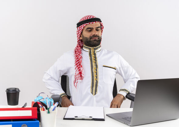 看身着传统服装的阿拉伯商人坐在桌旁 手持笔记本电脑 带着自信的表情在办公室工作阿拉伯语电脑表情