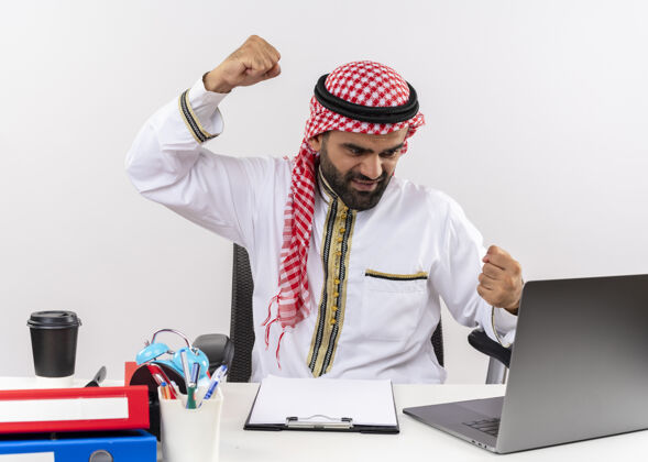 拳头身着传统服装的阿拉伯商人坐在桌旁 手提电脑紧握拳头 一脸愤怒地在办公室工作桌子传统穿