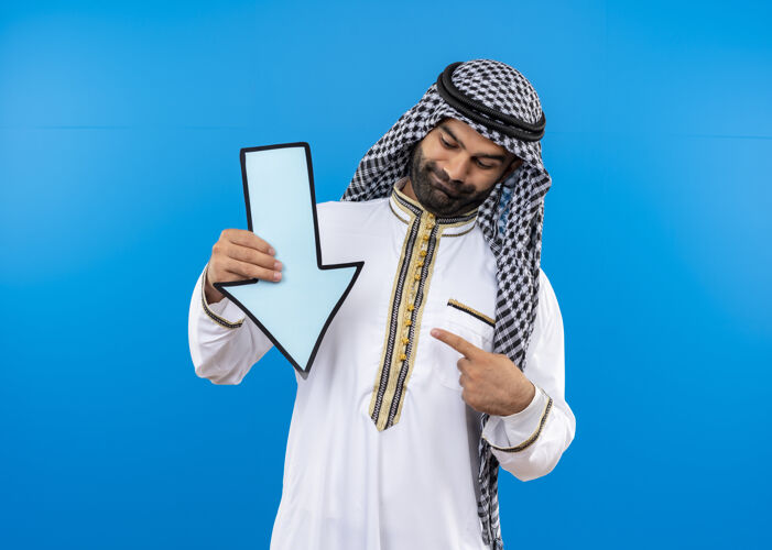手指身着传统服装的阿拉伯男子手持蓝色大箭头 用手指指着它微笑着站在蓝色的墙上指向传统向下