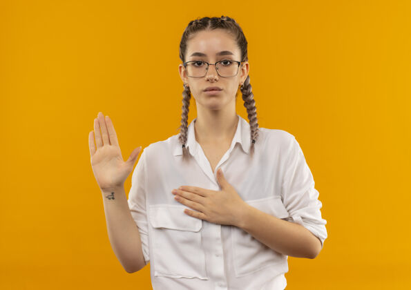 采取戴着眼镜 梳着辫子 穿着白衬衫的年轻女学生站在橙色的墙上 望着前面宣誓或许下诺言辫子制造衬衫