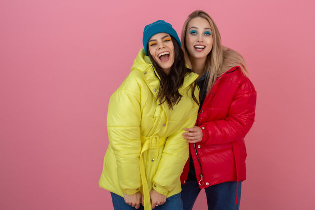 多彩两位迷人的活跃女性穿着鲜艳的红黄相间的冬季羽绒服在粉色的墙上摆着姿势 朋友们在一起玩得很开心 温暖的外套时尚潮流 疯狂搞笑的面孔微笑女士肖像