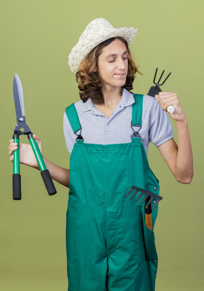 男人年轻的园丁穿着连体衣戴着帽子拿着树篱剪站着自信连身衣