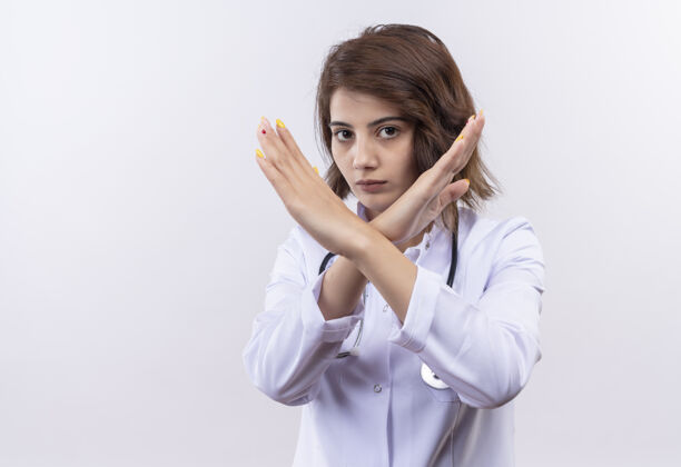 女人身穿白大褂 手持听诊器的年轻女医生做着自卫的手势 双臂交叉在脸上 站在白墙上站立外套交叉