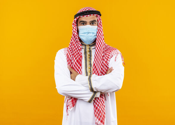 自信身着传统服装和面罩的阿拉伯商人自信地站在橙色的墙上面部保护面具