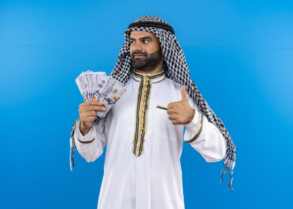 男人身着传统服装的阿拉伯男子站在蓝色的墙上 一边看一边微笑 一边自信地竖起大拇指传统拇指信心