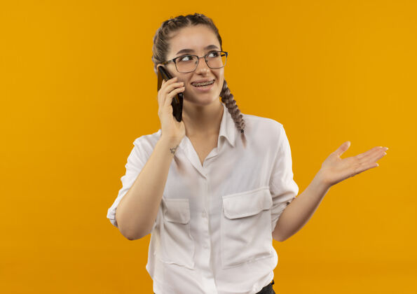 侧线戴着眼镜 梳着辫子 穿着白衬衫的年轻女学生在讲手机 微笑着用胳膊指着站在橙色墙上的一边指着女孩手势
