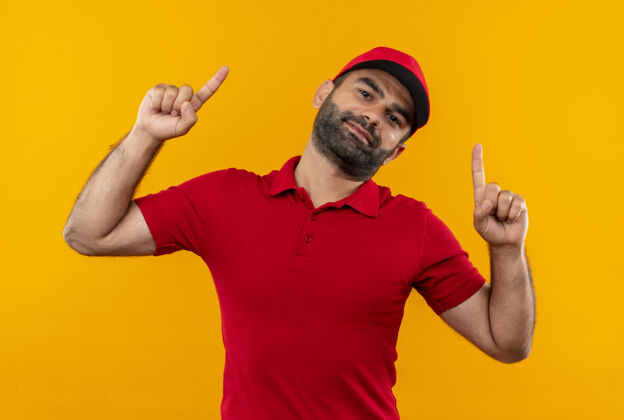 制服有胡子的送货员穿着红色制服 戴着帽子 食指朝上 微笑着站在橙色的墙上男人交货手指