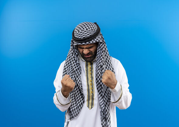 拳头身着传统服装的阿拉伯男子站在蓝色的墙上 一脸愤怒地握紧拳头传统脸男人