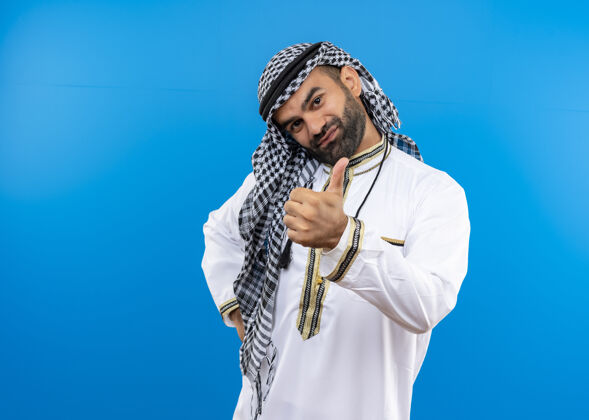 男人身着传统服装的阿拉伯男子站在蓝色的墙上 脸上带着微笑 竖起大拇指站秀微笑