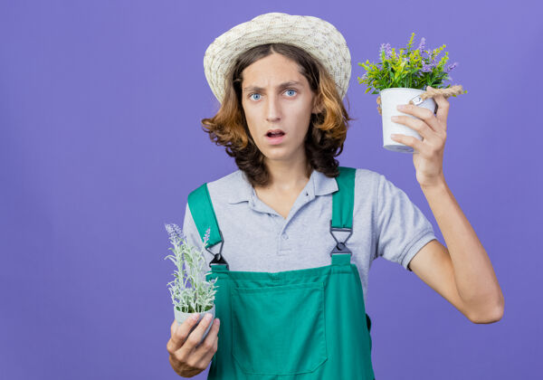 连身衣年轻的园丁穿着连体衣戴着帽子拿着盆栽植物被搞糊涂了帽子穿困惑