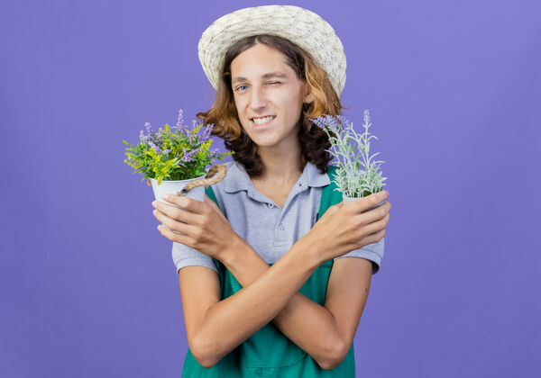 植物年轻的园丁穿着连体衣戴着帽子拿着盆栽微笑着年轻人帽子男人
