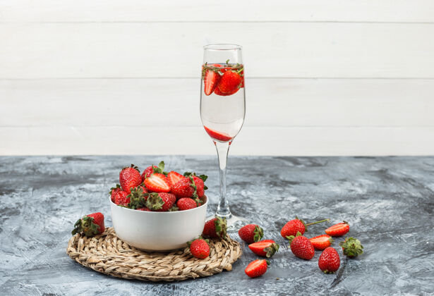 甜点把一碗草莓放在圆形柳条垫子上 一杯饮料放在深蓝色大理石和白色木板上水平餐垫玻璃生的