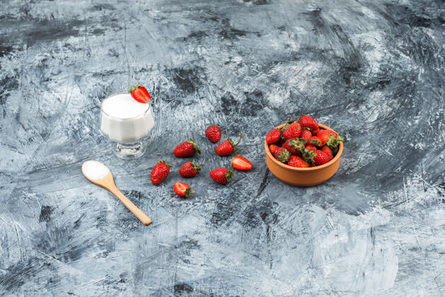 奶油俯视图：一杯酸奶放在柳条垫子上 木勺和草莓放在深蓝色大理石和白色木板上垂直牛奶草莓沙司