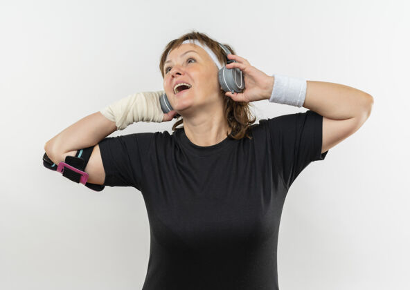 音乐中年运动女性 身穿黑色t恤 头戴头带 头戴耳机 站在白墙上 享受着她最喜爱的音乐快乐而积极的微笑站立耳机积极