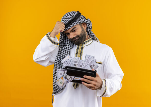 穿身着传统服饰的阿拉伯商人手里拿着钱包 手里拿着现金 站在橘色的墙上显得迷茫和焦急钱包持有焦虑