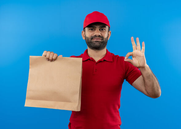包装有胡子的送货员穿着红色制服 戴着帽子 手里拿着纸包 微笑着 蓝色的墙上挂着“ok”的牌子胡子送货帽子