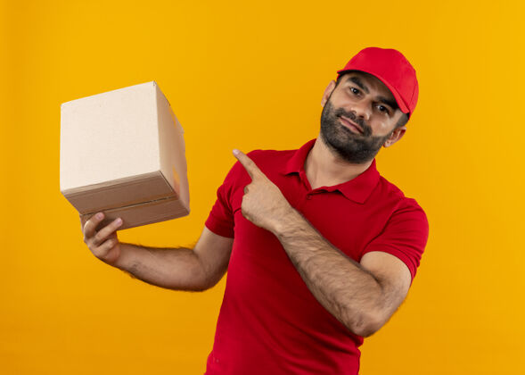 胡子有胡子的送货员穿着红色制服 拿着帽子的盒子 手指着包裹 微笑着自信地站在橙色的墙上指着送货拿着