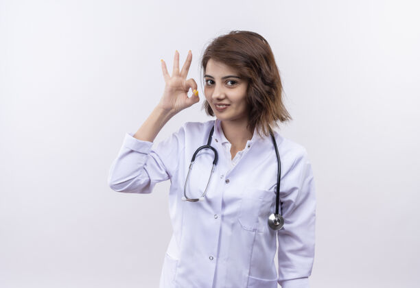 医生身穿白大褂 手持听诊器的年轻女医生微笑着 自信地站在白墙上做着“没事”的手势站着年轻外套