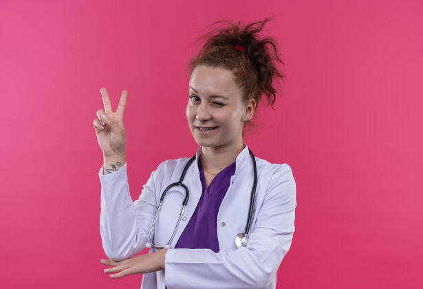 微笑年轻的女医生穿着白大褂 听诊器微笑着眨眼 粉红色的墙上立着胜利的标志医生展示胜利