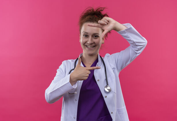 外套年轻的女医生穿着白色外套 戴着听诊器 手指微笑着 站在粉红色的墙上 愉快地看着这个框架穿站使