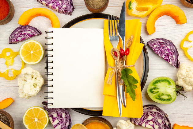 领带俯视食谱写在记事本上 把叉子和刀子绑在黄色餐巾上 把蔬菜切成圆形 把不同的香料放在碗里 放在白色的桌子上圆的餐巾顶部