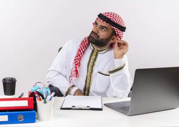 坐着身着传统服装的阿拉伯商人坐在桌子旁 拿着笔记本电脑一边看一边困惑地在办公室工作工作电脑商务人士