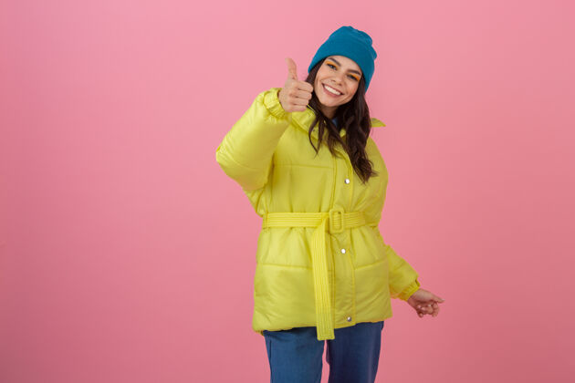 温暖迷人的微笑兴奋的时尚女人展示拇指在冬季时装造型在粉红色的墙上在明亮的霓虹灯黄色夹克 戴蓝色针织帽子 穿着暖和的衣服五颜六色羽绒服快乐