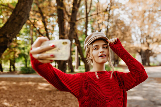 自拍美丽的金发女郎在秋季公园自拍穿着红毛衣戴白帽子的迷人女士拍照玩耍裙子户外