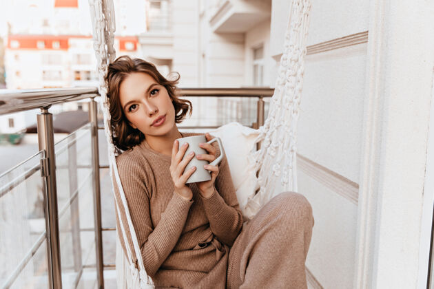 户外穿着针织连衣裙的漂亮女孩早上喝咖啡浪漫的白人年轻女子在阳台上端着一杯茶寒冷咖啡女人