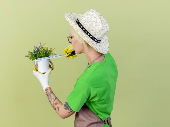 测量一位年轻的园丁 围裙上留着短发 戴着帽子 手里拿着盆栽植物 拿着卷尺 自信地站在明亮的背景上头发胶带举行