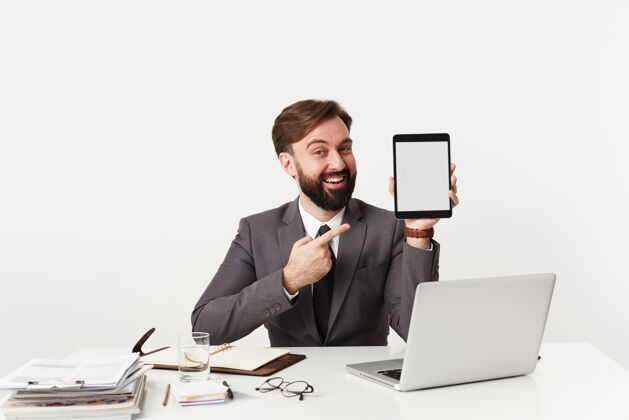 积极积极的年轻人 留着黑胡子 留着短发 手拿平板电脑 坐在白墙上的桌子旁 身穿灰色西装 面带微笑地看着前面办公室肖像姿势