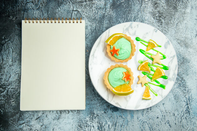 深色顶视图小馅饼与绿色糕点奶油和柠檬片在盘子上笔记本在黑暗的桌子上柠檬菜肴小馅饼