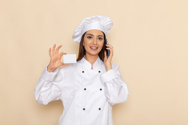 套装身穿白色厨师服的年轻女厨师手持名片 在浅白的墙上讲电话举行厨房交谈