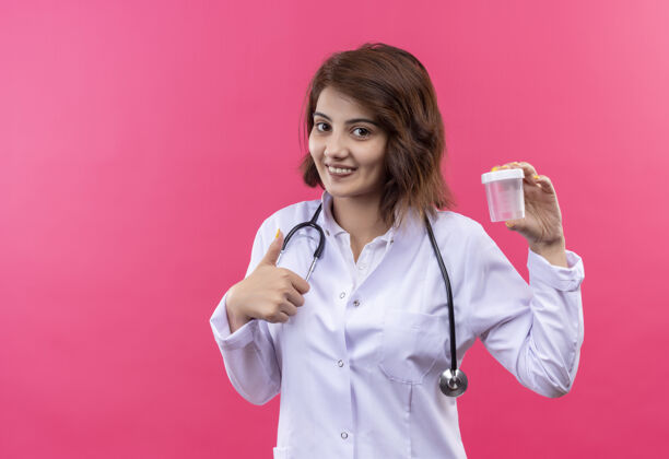 女人身穿白大褂的年轻女医生拿着听诊器 兴高采烈地笑着竖起大拇指听诊器展示微笑