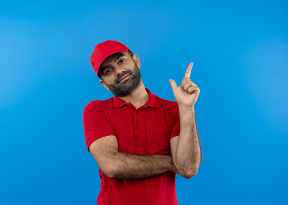 胡子留着胡须的送货员穿着红色制服 戴着帽子 手指指向一边 站在蓝色的墙上 看上去很自信手指壁板信心