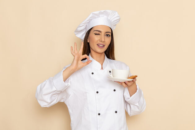 工作身穿白色厨师服的年轻女厨师正拿着一杯咖啡 在白墙上眨眼正面套装专业