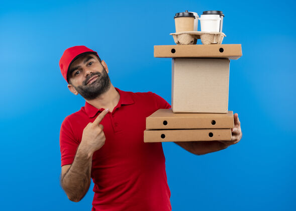 站着一个留着胡须 穿着红色制服 戴着帽子 拿着纸板箱的送货员站在蓝色的墙上 用手指着纸板箱 脸上带着微笑手指微笑拿着