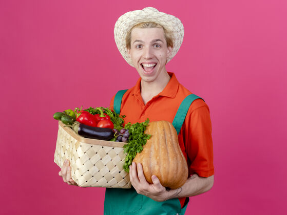 微笑年轻的园丁穿着连体衣戴着帽子拿着装满蔬菜的箱子蔬菜男人箱子