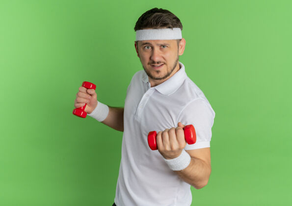 站立身穿白衬衫 头箍戴着哑铃的年轻健身男子站在绿色的墙上锻炼身体市民男人姿势