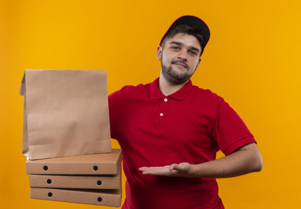 披萨年轻的送货员穿着红色制服 戴着帽子 手里拿着纸包和一叠比萨饼盒 看上去很自信立场制服男人