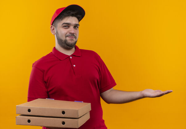 复制年轻的送货员身穿红色制服 头戴鸭舌帽 手里拿着一叠比萨饼盒 用手臂哦 他的手抄写空间年轻人哦盒子