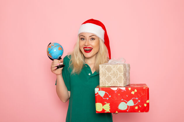 地球仪正面图年轻女性手持地球仪和圣诞礼物在粉红色墙上的照片模特妇女圣诞节新年彩色假期微笑年轻女性颜色