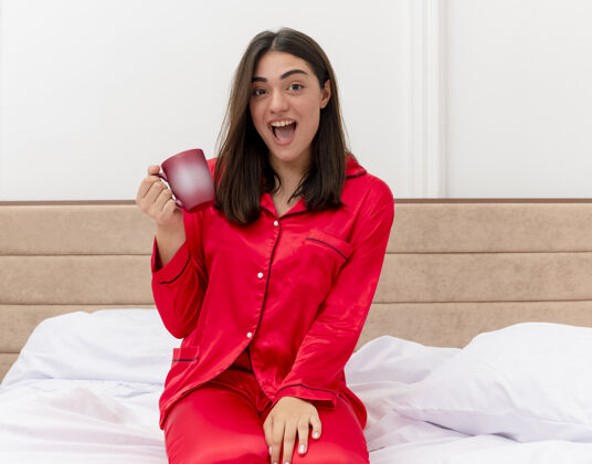 杯子穿着红色睡衣的快乐年轻美女坐在床上 端着一杯咖啡 看着相机 在卧室的室内灯光背景下 快乐而兴奋红色快乐室内