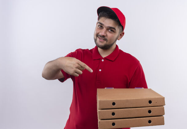 站着年轻的送货员穿着红色制服 戴着帽子 拿着披萨盒 手指着披萨盒 自信地微笑着微笑帽子男人