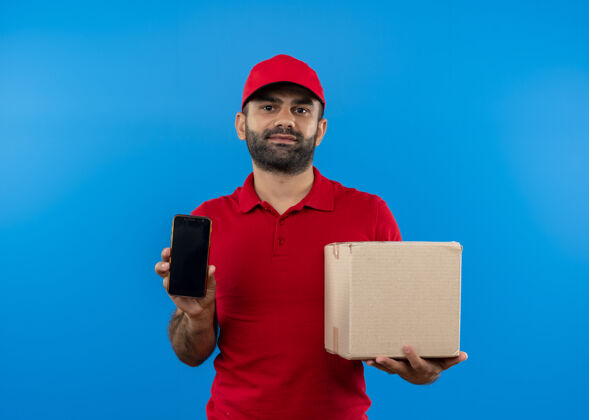 站着留着胡须的送货员身穿红色制服 戴着帽子 手里拿着打开的披萨盒 在蓝色的墙上展示着表情自信的智能手机打开移动帽子