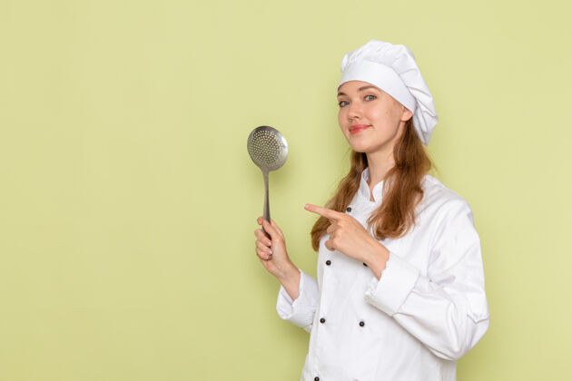 持有身穿白色厨师服的女厨师手持大银勺站在绿色墙上的正视图专业前面穿